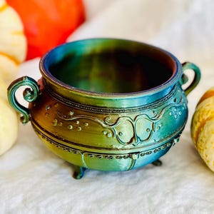 May include: A teal and gold metallic cauldron with decorative detailing and small handles. The cauldron is set against a white fabric backdrop, with orange, white, and yellow pumpkins surrounding it.