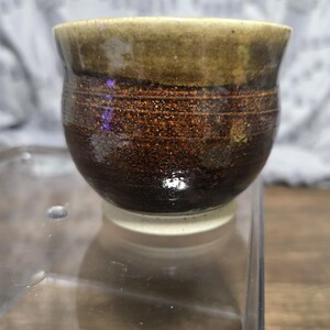 May include: A handmade ceramic cup with a rounded base and a slightly flared rim. The cup features a gradient of colors, transitioning from a light tan rim to a dark brown body. The base is a clean white. The cup is likely used for drinking.