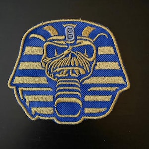 Puede incluir: Parche bordado con un diseño azul y dorado de una máscara de faraón. El parche tiene un aspecto detallado y texturizado con rayas y acentos horizontales dorados. La imagen central es una calavera estilizada con una cofia, sobre un fondo oscuro.