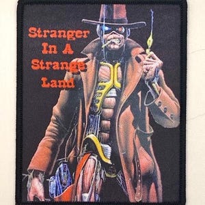 Pode incluir: Um patch preto com uma ilustração detalhada de uma figura com casaco e chapéu castanhos, segurando um pequeno objeto. O texto "Stranger In A Strange Land" está em letras vermelhas.
