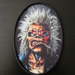 Könnte beinhalten: Ovaler Aufnäher mit einer detaillierten Illustration einer skelettartigen Figur mit weißem Haar, roten und blauen Akzenten und einer Zigarette. Der Aufnäher hat einen schwarzen Rand, für Kleidung oder Accessoires.