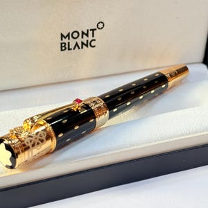 K&ouml;nnte beinhalten: Ein schwarzer und goldener Montblanc-Stift mit gepunktetem Muster, in seiner Markenschachtel. Der Stift hat goldene Akzente, einen roten Edelstein und das Montblanc-Logo. Die Box ist schwarz mit wei&szlig;em Innenraum.