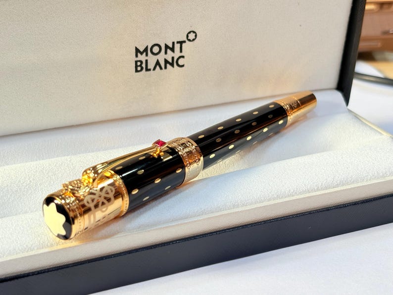 K&ouml;nnte beinhalten: Ein schwarzer und goldener Montblanc-Stift mit gepunktetem Muster, in seiner Markenschachtel. Der Stift hat goldene Akzente, einen roten Edelstein und das Montblanc-Logo. Die Box ist schwarz mit wei&szlig;em Innenraum.