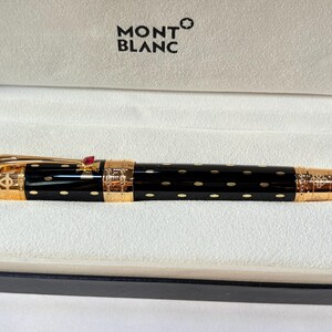 K&ouml;nnte beinhalten: Ein schwarzer Montblanc-Stift mit goldenen Akzenten und einem roten Edelstein. Der Stift hat ein gepunktetes Muster und wird in einer wei&szlig;en Box mit dem Montblanc-Logo pr&auml;sentiert.