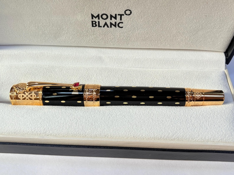 K&ouml;nnte beinhalten: Ein schwarzer Montblanc-Stift mit goldenen Akzenten und einem roten Edelstein. Der Stift hat ein gepunktetes Muster und wird in einer wei&szlig;en Box mit dem Montblanc-Logo pr&auml;sentiert.