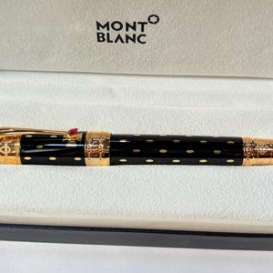 K&ouml;nnte beinhalten: Ein schwarzer Montblanc-Stift mit goldenen Akzenten und einem roten Juwel. Der Stift hat ein gepunktetes Muster und wird in einer wei&szlig;en Schachtel mit dem Montblanc-Logo pr&auml;sentiert.