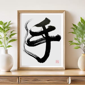 以下が含まれることがあります： 黒と白の額入り日本の書道アートプリント。アートワークは、大胆で表現力豊かな筆致で文字を形成しています。プリントは木製の棚に飾られており、白とタンの花瓶に入った緑の植物が両側にあります。