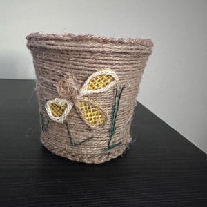 Può includere: Un piccolo vaso cilindrico avvolto in spago color naturale. Un fiore decorativo fatto di spago e rete gialla è attaccato sul davanti. Accenti di spago verde aggiungono un tocco di colore.