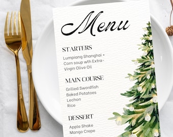 May include: A white menu card with a watercolor Christmas tree design. The menu includes starters, main courses, and desserts. Gold-colored fork and knife are on the left side of the plate. The menu items are listed in black text.