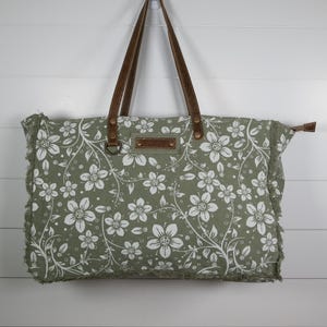 May include: A rectangular tote bag with a floral pattern in white on a muted green background. The bag has brown leather handles and a brown leather patch. The edges of the bag are frayed.