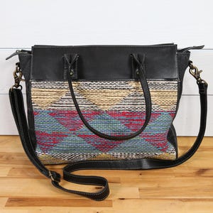 May include: Black leather tote bag with a woven fabric front panel. The fabric features a geometric pattern in red, blue, and tan. The bag has two handles and a detachable shoulder strap. The bag is sitting on a wooden surface.