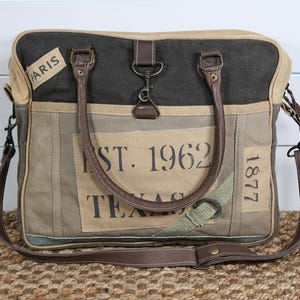 May include: Canvas tote bag with brown leather handles and a detachable shoulder strap. The bag features the text "EST. 1962 TEXAS" and "PARIS" on a tan background, with a dark gray top and accents.