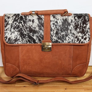 May include: A brown leather satchel with a cowhide flap. The bag has a top handle, a shoulder strap, and a brass clasp. The cowhide flap is black and white. The bag is sitting on a wooden surface.