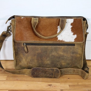 May include: A brown leather messenger bag featuring a cowhide flap with brown and white patches. The bag includes two top handles, a detachable shoulder strap with a padded section, and a horizontal zippered pocket on the front exterior.