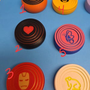 Puede incluir: Una colección de posavasos redondos y coloridos con varios diseños. Los posavasos presentan imágenes de una calavera, un corazón, una máscara de Iron Man, un gato, una mariposa y un conejo. Los posavasos son en tonos naranja, negro, rojo, amarillo, rosa, azul y crema.