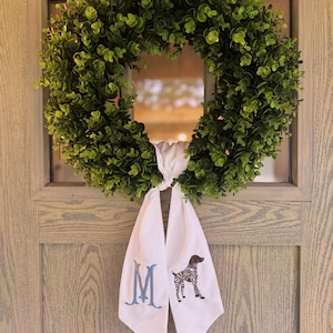 May include: A green leafy wreath hangs on a wooden door. A white fabric scarf is tied to the wreath, embroidered with a blue letter "M" and a brown and white dog illustration.