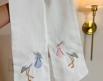 May include: Two white fabric items with embroidered storks carrying bundles. One stork holds a blue bundle, the other a pink one. The storks have gray wings and yellow legs. The items are likely baby-themed.