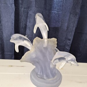 May include: A frosted glass sculpture featuring three dolphins leaping from a wave-like base. The dolphins are depicted in mid-air, with their bodies gracefully arched. The sculpture has a light blue hue and sits on a round base.