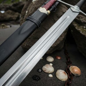 Puede incluir: Una espada de acero de doble filo con un canal central, que presenta una guarda y una vaina envuelta en cuero negro con una sección de empuñadura de color rojo oscuro y detalles metálicos, colocada en diagonal con una hoja pulida.