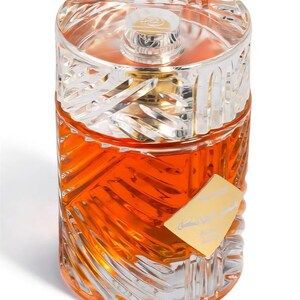 May include: A clear glass perfume bottle with an orange liquid inside. The bottle has a silver-colored, geometric-patterned cap and a gold-colored label. The bottle's design features a repeating, etched pattern.