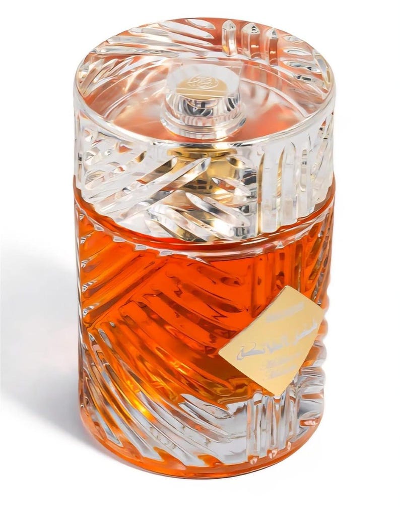 May include: A clear glass perfume bottle with an orange liquid inside. The bottle has a silver-colored, geometric-patterned cap and a gold-colored label. The bottle's design features a repeating, etched pattern.