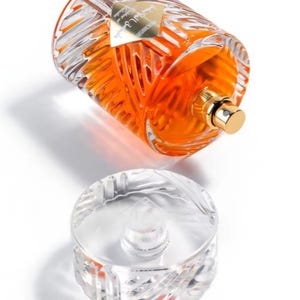 May include: A clear glass perfume bottle with an orange liquid inside, lying on its side. The bottle has a gold cap and a decorative, textured design. The clear, detached stopper is below the bottle.