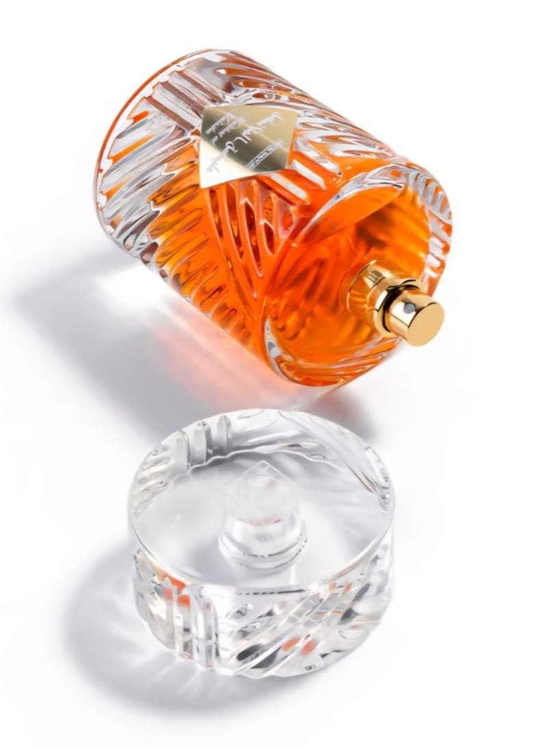 May include: A clear glass perfume bottle with an orange liquid inside, lying on its side. The bottle has a gold cap and a decorative, textured design. The clear, detached stopper is below the bottle.