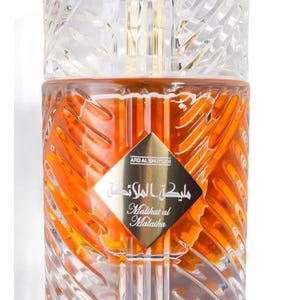 May include: A perfume bottle with a clear, textured cap and a golden interior. The bottle has a diamond-shaped label with Arabic script and the English translation "Malikat al Malaika". The liquid inside is orange.
