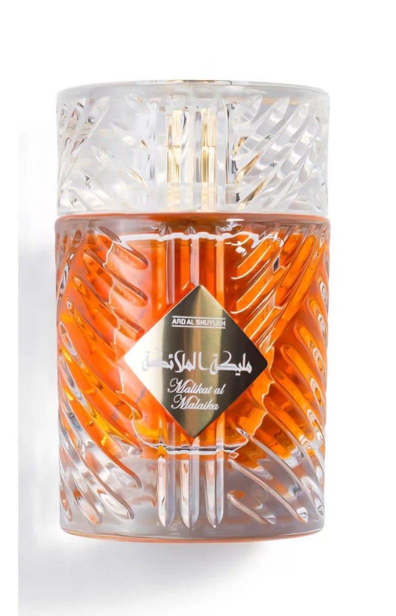 May include: A perfume bottle with a clear, textured cap and a golden interior. The bottle has a diamond-shaped label with Arabic script and the English translation "Malikat al Malaika". The liquid inside is orange.