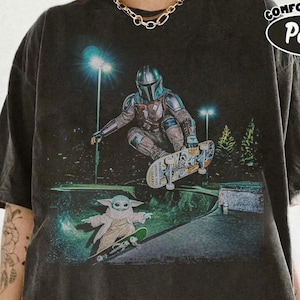 Op de afbeelding: Donkergrijs t-shirt met een afbeelding van The Mandalorian en Grogu die skaten in een skatepark. Het ontwerp bevat een nachtsc&egrave;ne met groene en blauwe accenten. De tekst "COMFORT COLORS Pepper" staat rechtsboven.