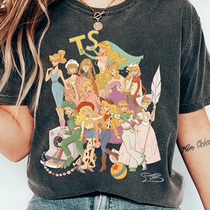 Puede incluir: Una camiseta de manga corta gris carb&oacute;n con un estampado gr&aacute;fico de varios personajes animados con diversos atuendos. El dise&ntilde;o incluye el texto amarillo TS en la parte superior, una c&aacute;mara vintage y una pelota colorida.