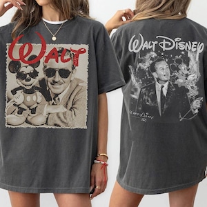 May include: A charcoal gray short-sleeve t-shirt featuring a front graphic of Walt Disney and Mickey Mouse wearing sunglasses with the red text Walt. The back displays a black and white graphic of Walt Disney and Mickey Mouse with the text Walt Disney.