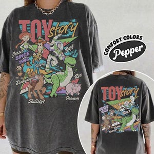 May include: A charcoal gray oversized t-shirt featuring a vintage-style Toy Story graphic print. The design includes characters Jessie, Woody, Buzz Lightyear, Slinky Dog, Bullseye, Rex, and Hamm with the text Toy Story and Since 1995.