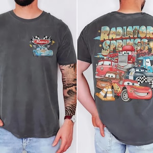 Puede incluir: Una camiseta gris carb&oacute;n de manga corta con un peque&ntilde;o gr&aacute;fico en el pecho que muestra un coche azul y el texto Name. La parte trasera presenta un gr&aacute;fico grande de Radiator Springs con varios coches de dibujos, un trofeo y conos.