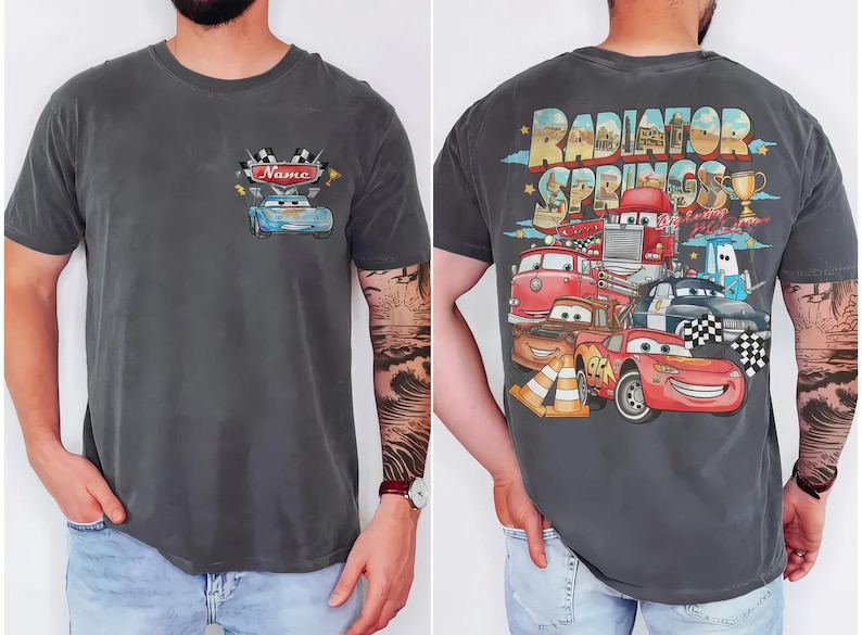 Puede incluir: Una camiseta gris carb&oacute;n de manga corta con un peque&ntilde;o gr&aacute;fico en el pecho que muestra un coche azul y el texto Name. La parte trasera presenta un gr&aacute;fico grande de Radiator Springs con varios coches de dibujos, un trofeo y conos.
