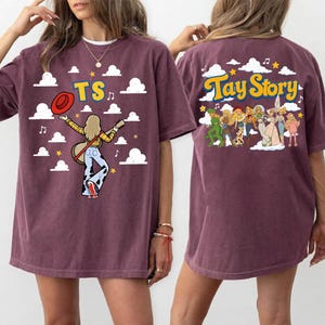 May include: A maroon short-sleeve t-shirt featuring a graphic print. The front displays the text TS with a figure holding a guitar and hat against a cloud background. The back shows a group of figures with the text Tay Story and clouds.