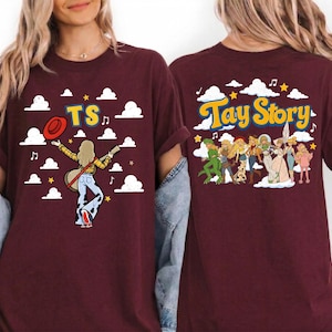 May include: A maroon t-shirt featuring a graphic print. The front displays a figure with a guitar, a red hat, and the text TS against a sky with clouds. The back shows a group of illustrated characters and the text Tay Story with clouds.