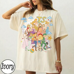 May include: An ivory t-shirt featuring a large graphic print of various animated characters in a stylized collage. The design includes the letters TS in yellow, blue clouds, a rocket, a sheriff star, and a camera. The characters are depicted in colorful outfits, including dresses, cowgirl attire, and space-themed clothing.