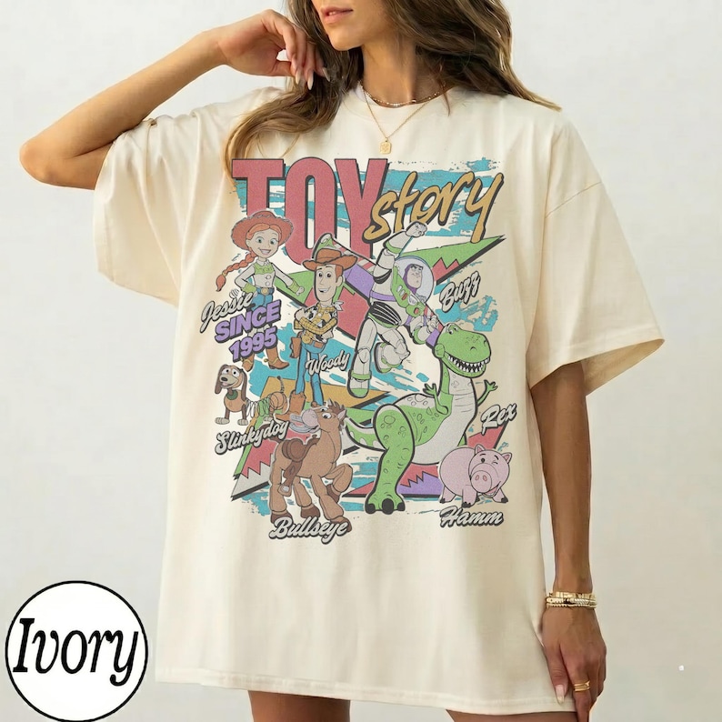 May include: An ivory short-sleeve t-shirt featuring a large graphic print of Toy Story characters including Jessie, Woody, Buzz Lightyear, Slinky Dog, Bullseye, Rex, and Hamm. The text reads Toy Story, Jessie Since 1995, Woody, Buzz, Slinky Dog, Bullseye, and Hamm.