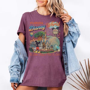 May include: A purple short-sleeve t-shirt featuring a graphic print of Florida Disney World. The design includes Mickey Mouse, the castle, the Epcot sphere, the Hollywood Tower, and the text Florida Disney World The Most Magical Place On Earth.