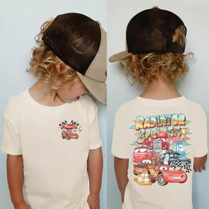 Puede incluir: Una camiseta color crema con un peque&ntilde;o gr&aacute;fico de un coche de carreras rojo y banderas a cuadros en el pecho izquierdo, y un gr&aacute;fico grande en la espalda con el texto Radiator Springs, Lightning McQueen y varios personajes de coches.