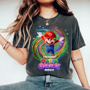 May include: A charcoal gray short-sleeve t-shirt featuring a graphic of Mario from the Super Mario Galaxy Movie. The character is centered against a colorful, swirling cosmic background with stars and planets. The text reads THE SUPER MARIO GALAXY MOVIE.