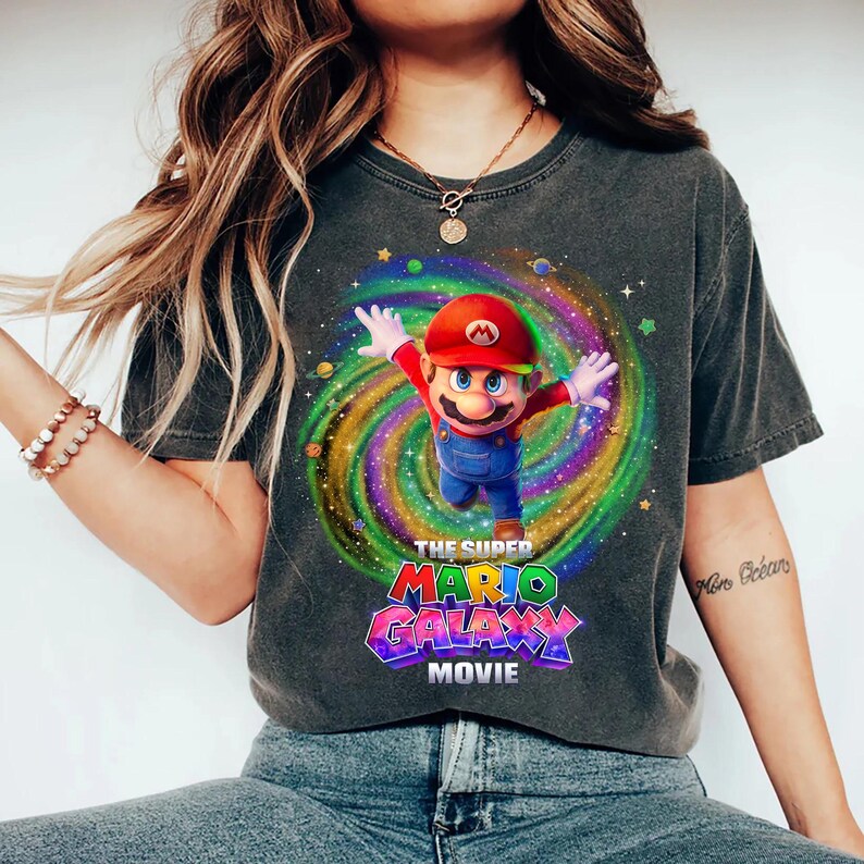 May include: A charcoal gray short-sleeve t-shirt featuring a graphic of Mario from the Super Mario Galaxy Movie. The character is centered against a colorful, swirling cosmic background with stars and planets. The text reads THE SUPER MARIO GALAXY MOVIE.