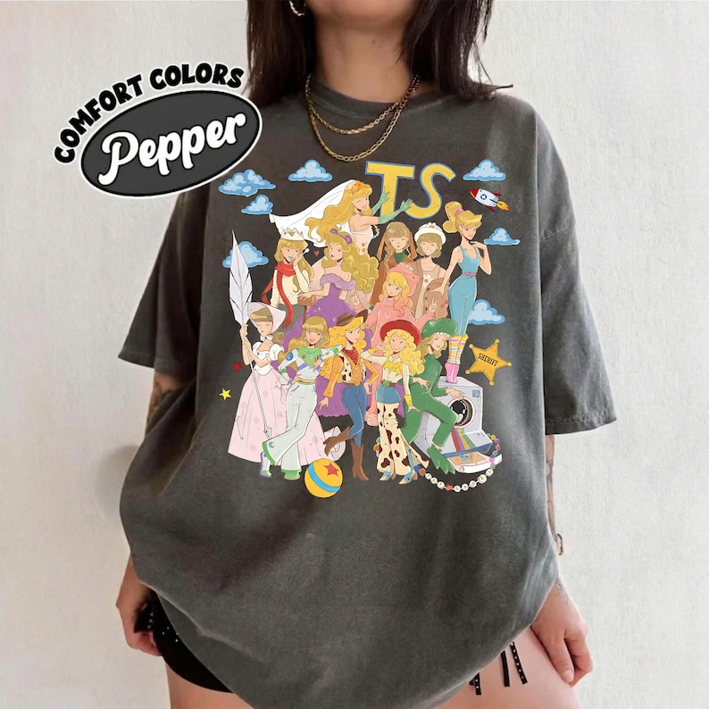 May include: A charcoal gray short-sleeve t-shirt featuring a graphic print of multiple animated characters in various outfits, including a yellow TS logo, a rocket, and a sheriff star. The text Comfort Colors Pepper is printed in the upper left corner.