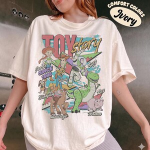May include: An ivory short-sleeve t-shirt featuring a graphic print of Toy Story characters including Jessie, Woody, Buzz Lightyear, Slinky Dog, Bullseye, Rex, and Hamm. The text reads Toy Story, Since 1995, Jessie, Woody, Buzz, Slinky Dog, Bullseye, Rex, and Hamm.