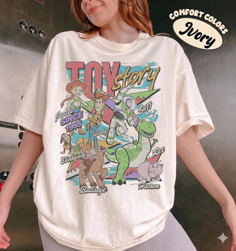 May include: An ivory short-sleeve t-shirt featuring a graphic print of Toy Story characters including Jessie, Woody, Buzz Lightyear, Slinky Dog, Bullseye, Rex, and Hamm. The text reads Toy Story, Since 1995, Jessie, Woody, Buzz, Slinky Dog, Bullseye, Rex, and Hamm.
