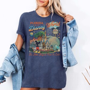 May include: A dark blue heathered t-shirt featuring a colorful graphic print of Disney World landmarks, including Cinderella Castle, Epcot, and Mickey Mouse. The text reads Florida, Disney World, Est. 1971, and The Most Magical Place On Earth.