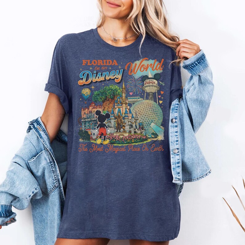 May include: A dark blue heathered t-shirt featuring a colorful graphic print of Disney World landmarks, including Cinderella Castle, Epcot, and Mickey Mouse. The text reads Florida, Disney World, Est. 1971, and The Most Magical Place On Earth.