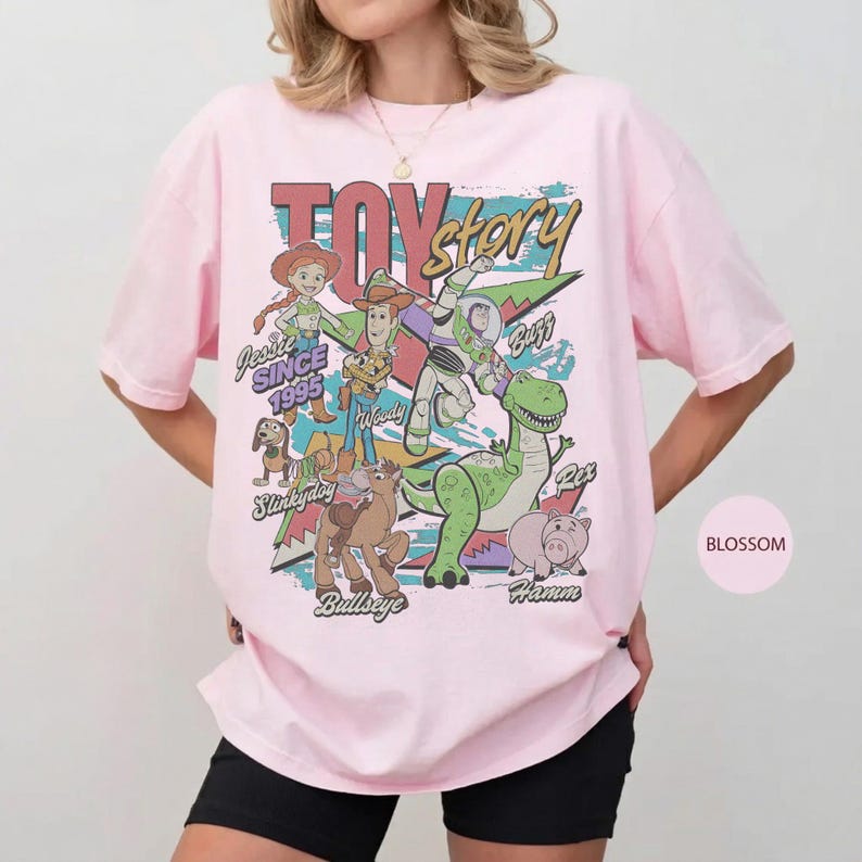 May include: A light pink t-shirt featuring a colorful graphic print of Toy Story characters including Jessie, Woody, Buzz Lightyear, Slinky Dog, Bullseye, Rex, and Hamm. The text reads TOY story, SINCE 1995, Jessie, Woody, Buzz, Slinky Dog, Bullseye, and Hamm.