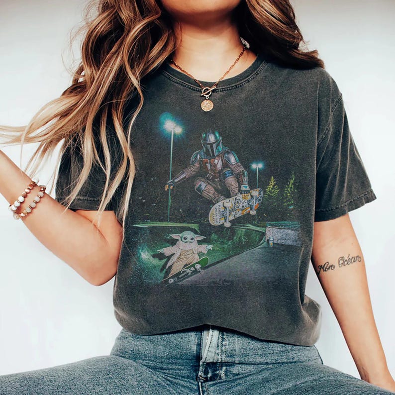 Op de afbeelding: Donkergrijs t-shirt met een afbeelding van The Mandalorian en Grogu die skateboarden in een skatepark. Het ontwerp bevat een gedetailleerde illustratie van de personages en skateboards, tegen een achtergrond van straatverlichting en bomen.